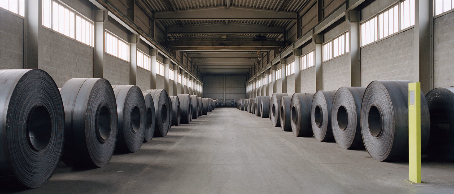 Rows of hot-rolled steel coils standing on end down the aisle of a storage hall.
