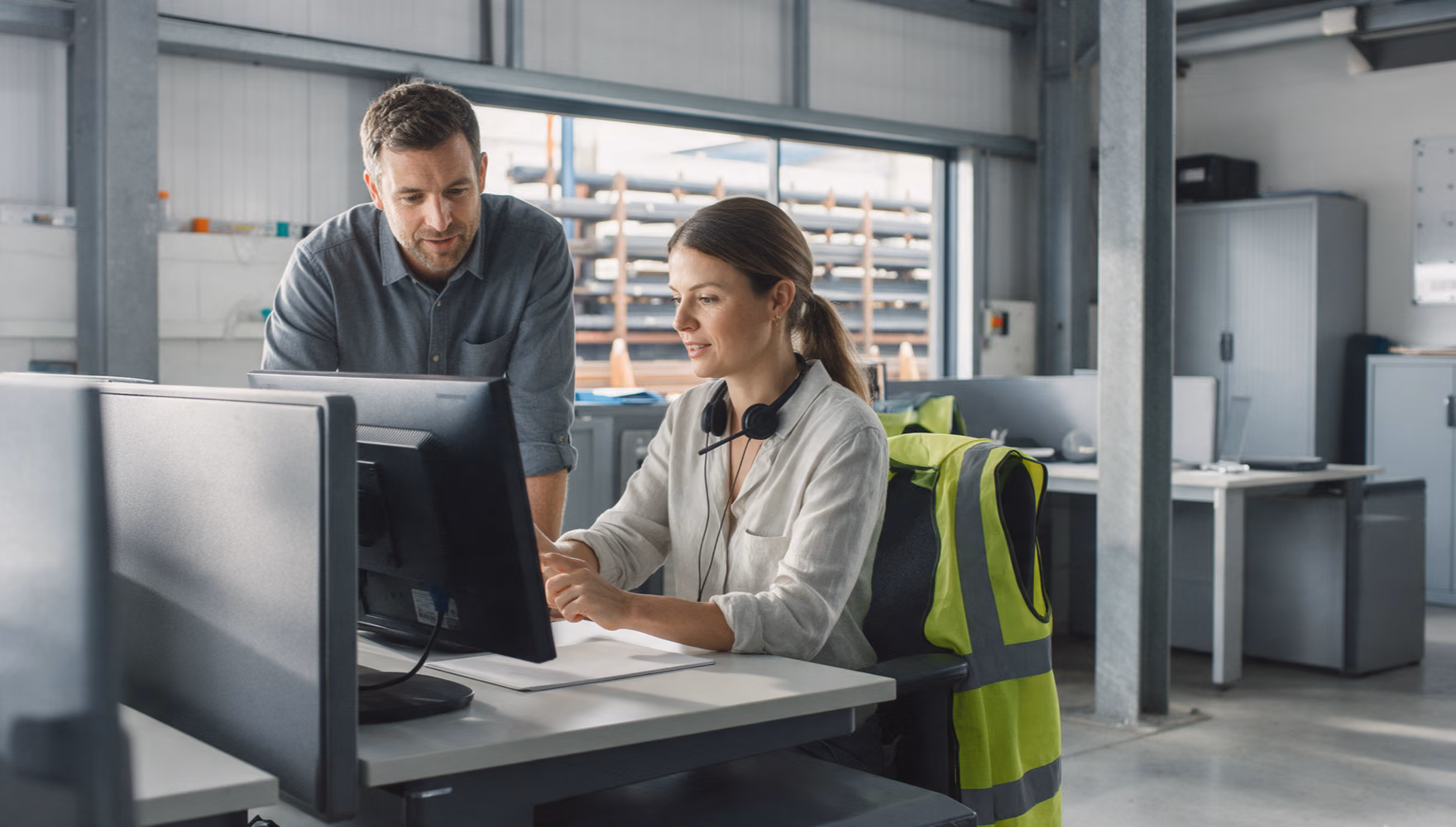 A woman with a headset around her neck turning her monitor towards a colleague at her desk in a sales office above a steel yard.