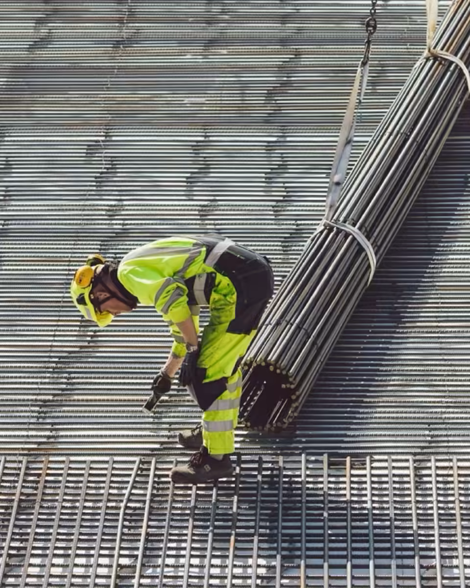 Ein Arbeiter in Warnkleidung führt ein Bündel Bewehrungsstäbe auf ein verlegtes Stahlgitter herab.