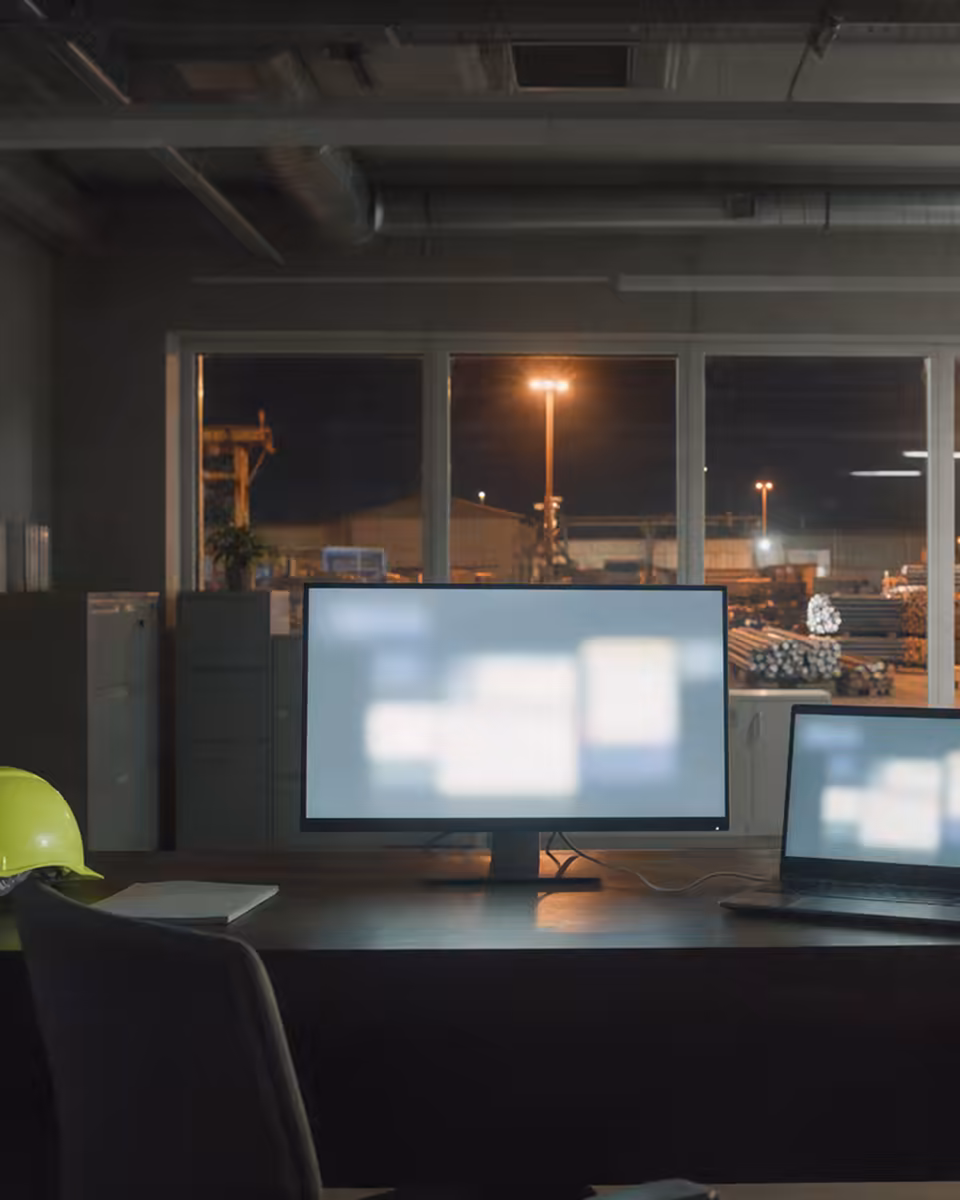 Ein Monitor und ein angedockter Laptop leuchten nachts auf einem Schreibtisch, dahinter das beleuchtete Stahllager vor dem Fenster.