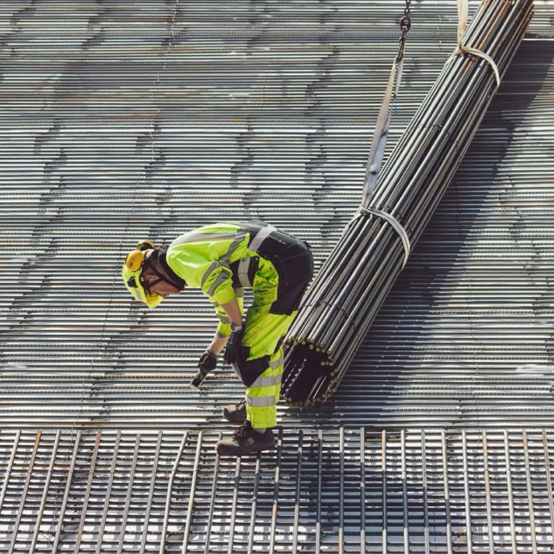 A worker in hi-vis clothing guiding a crane-slung bundle of reinforcement bars down onto a laid steel mesh.