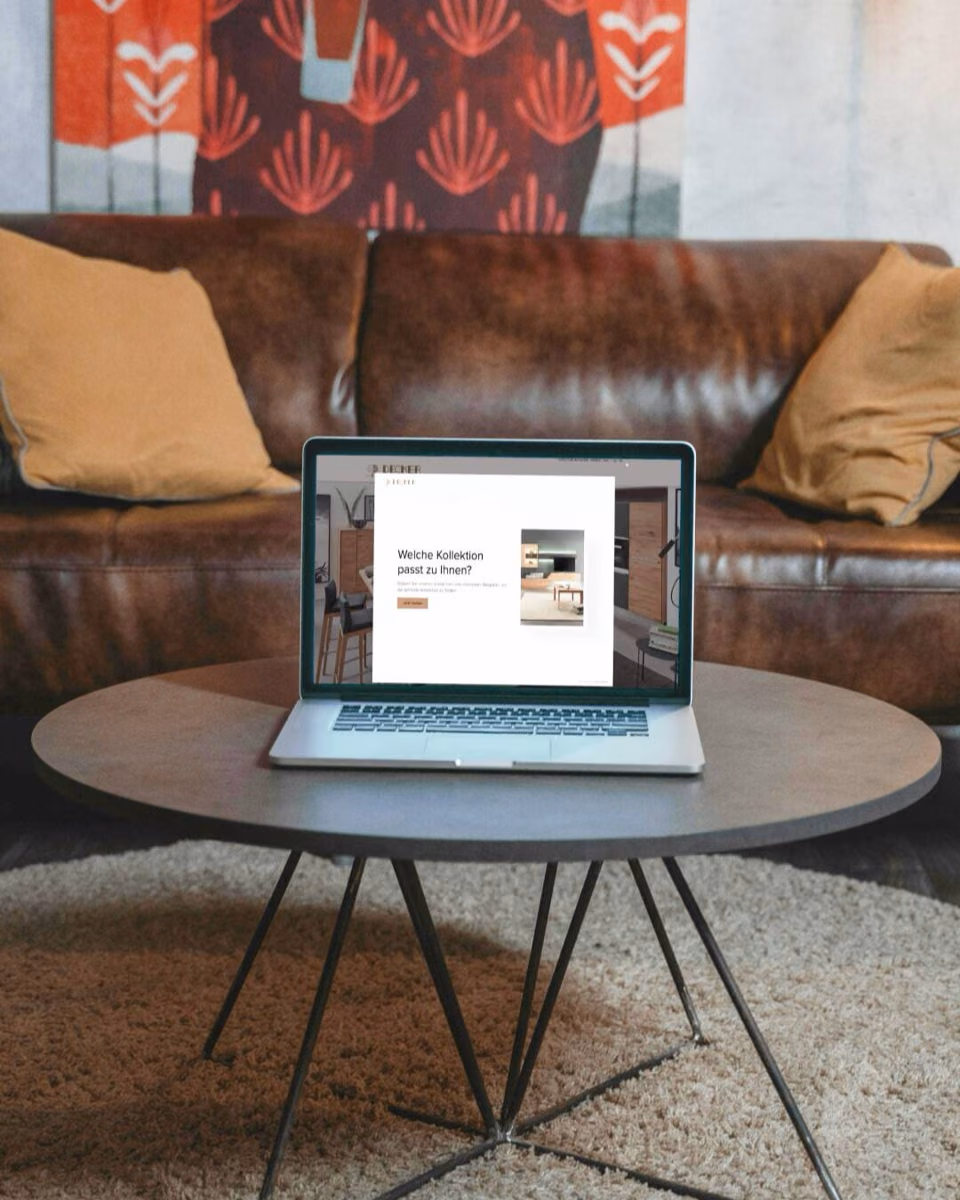 A laptop on a living room coffee table shows a pop-up asking which furniture collection suits the visitor.
