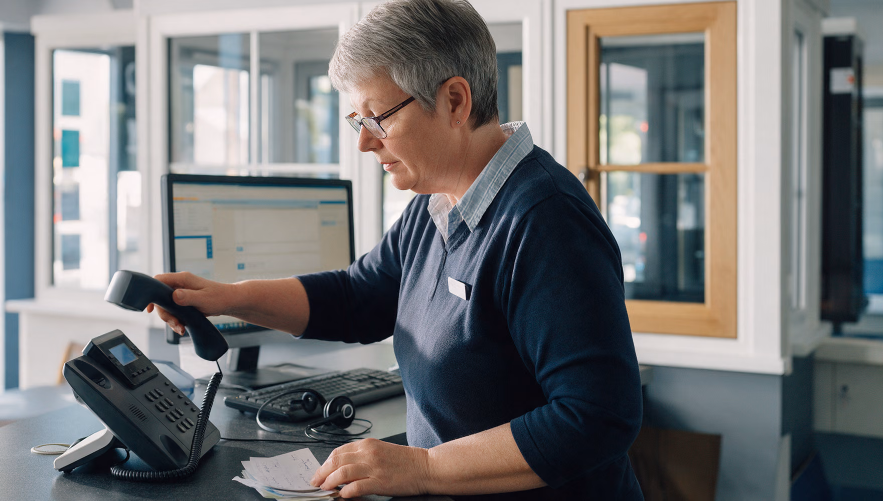 A customer adviser at the enquiries desk of a windows showroom lifting the desk phone handset while reading a stack of handwritten message slips.