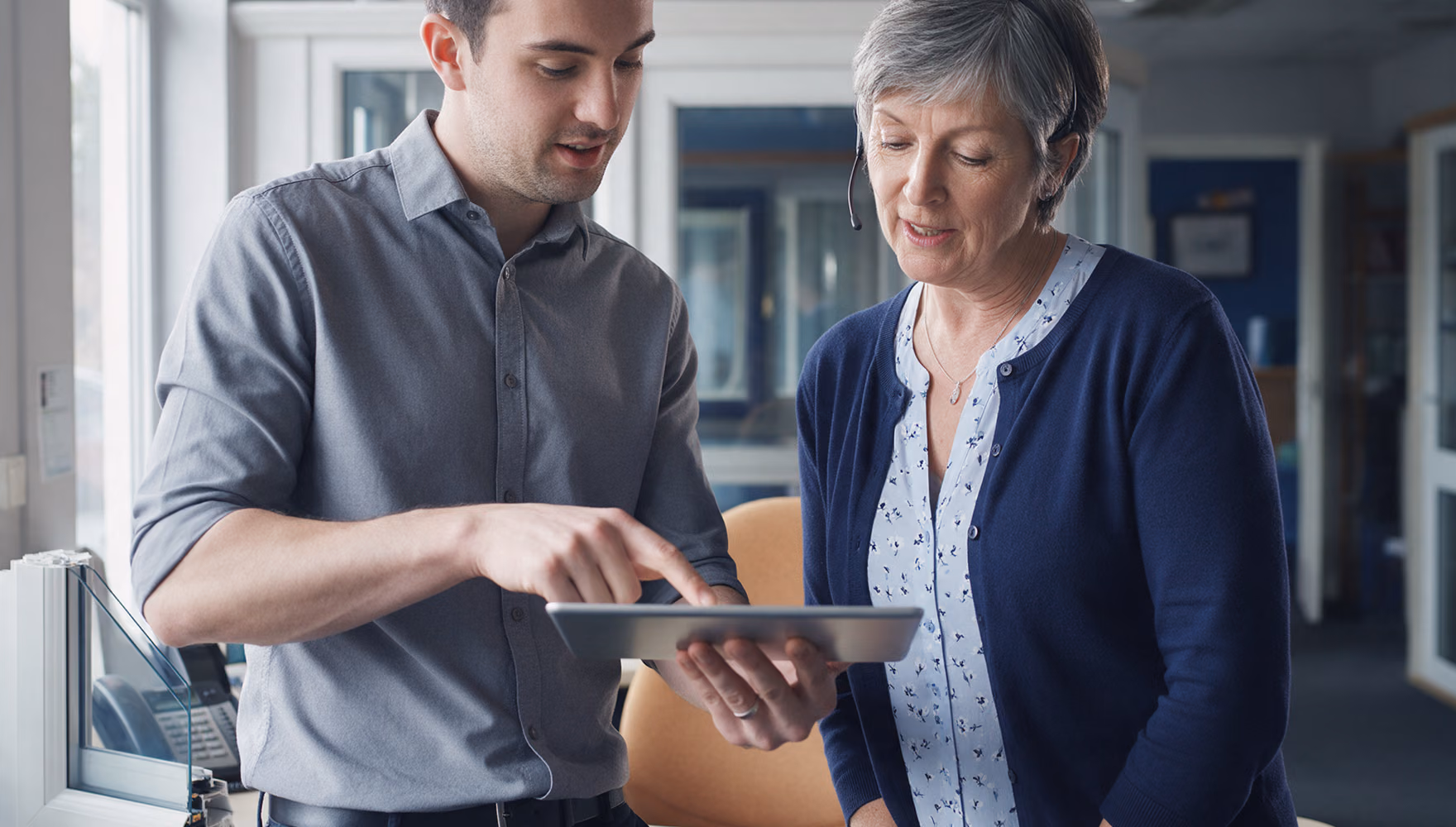 Two colleagues in the back office of a windows company reading an escalated enquiry together on a tablet, one of them wearing a headset.