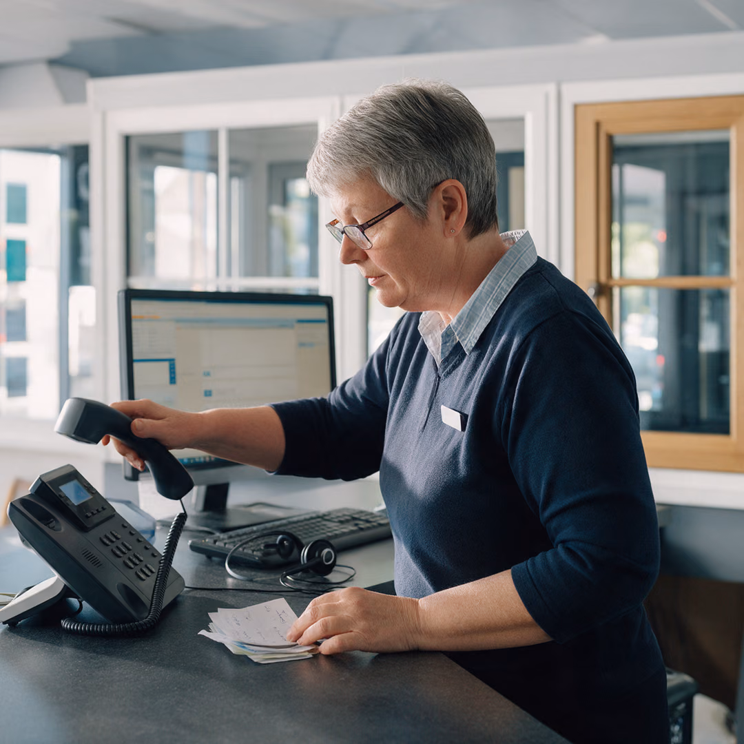 A customer adviser at the enquiries desk of a windows showroom lifting the desk phone handset while reading a stack of handwritten message slips.