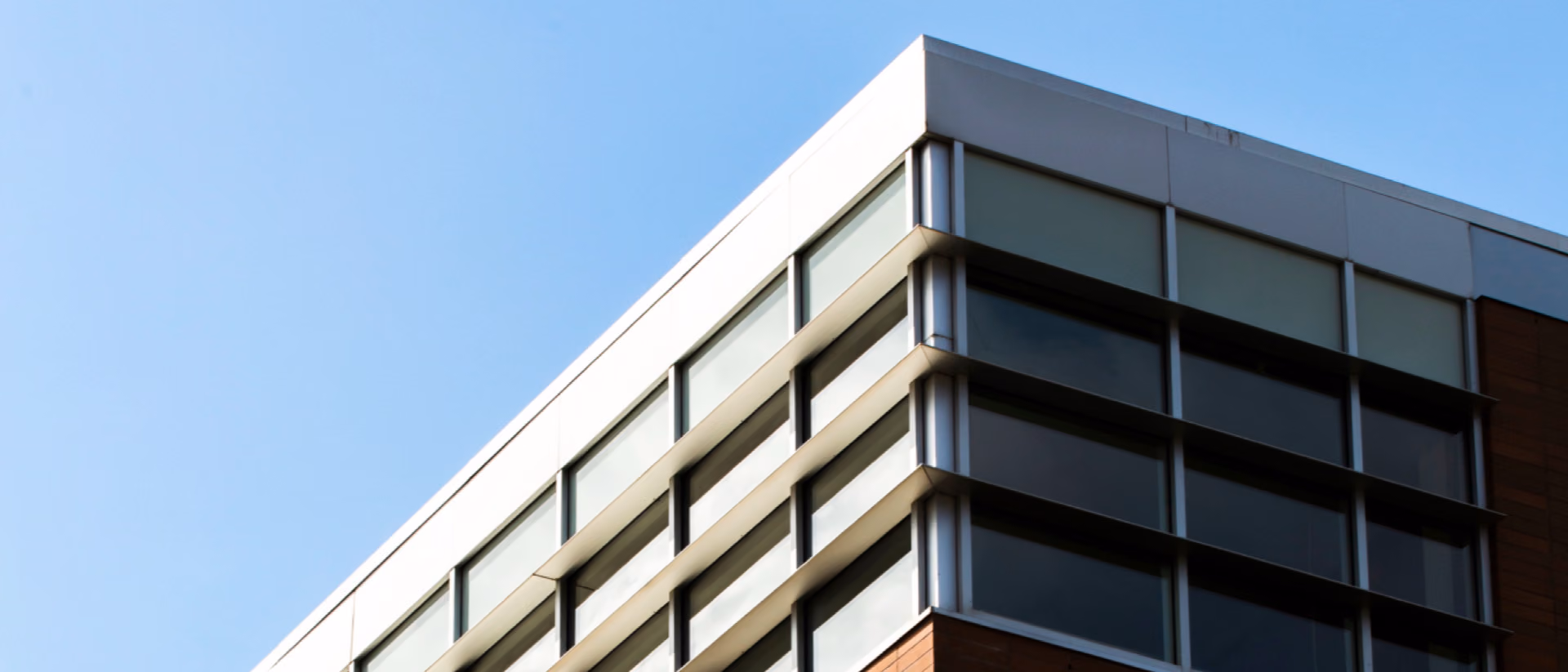 The top corner of an office building with banded glazing and brick cladding cuts into a cloudless blue sky.