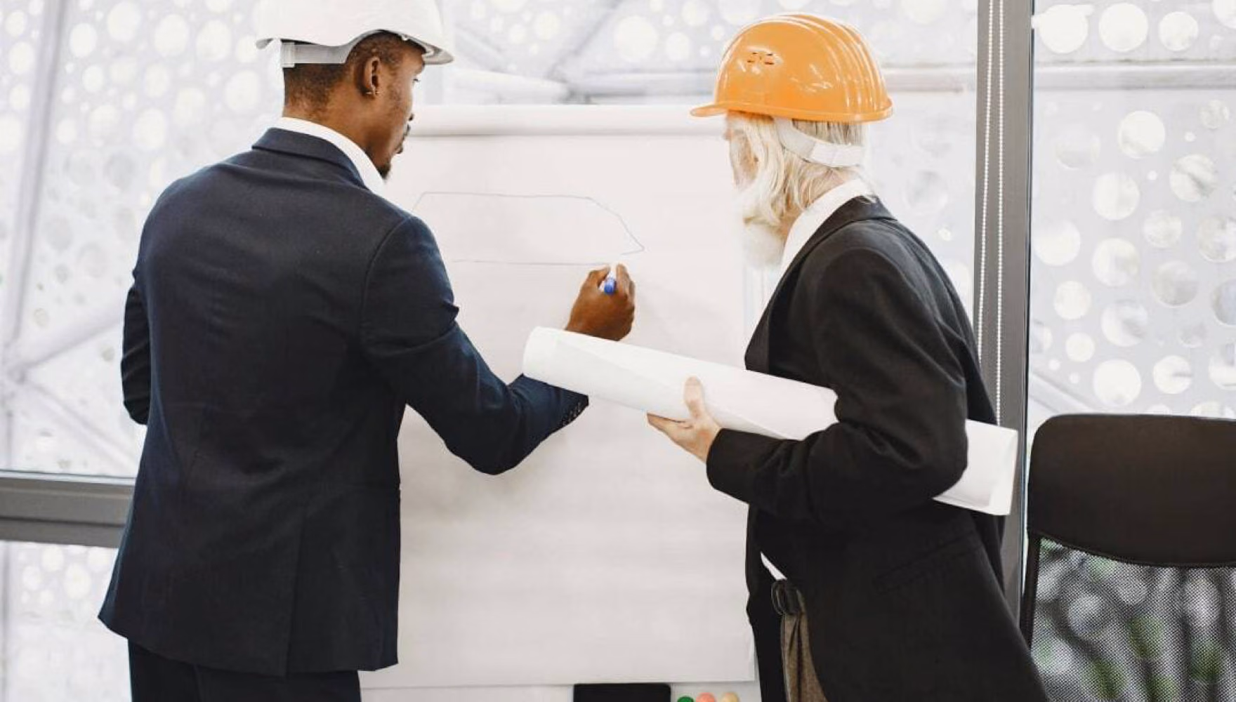 Two men in hard hats work at a whiteboard, one drawing while the other holds a rolled plan.