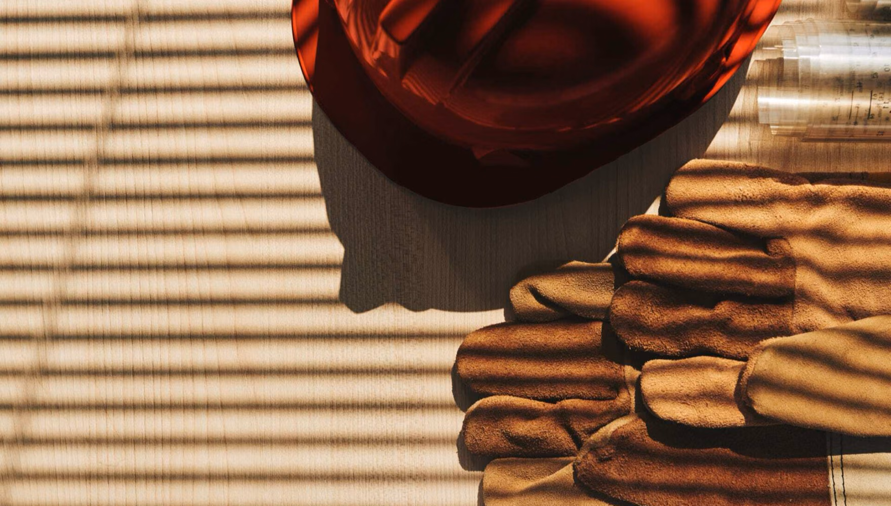 A red hard hat, leather work gloves and rolled-up plans lie ready on a slatted wooden surface.