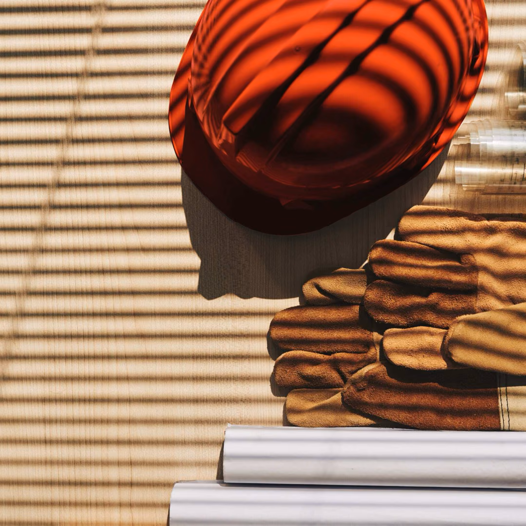 A red hard hat, a stack of leather work gloves and rolled-up plans lie on a slatted wooden surface.
