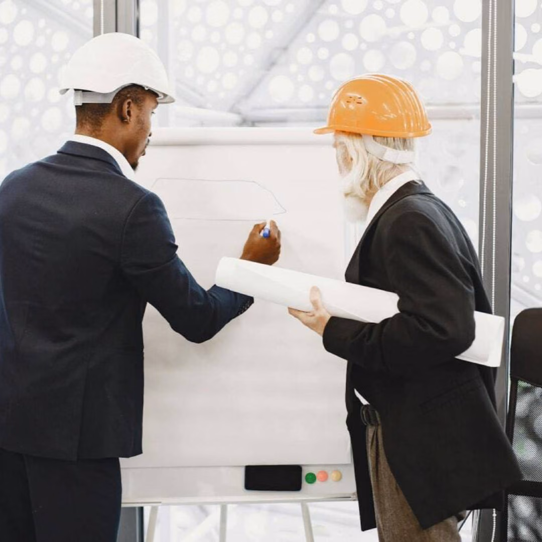 Two men in hard hats stand at a whiteboard, one drawing on it while the other holds a rolled plan.