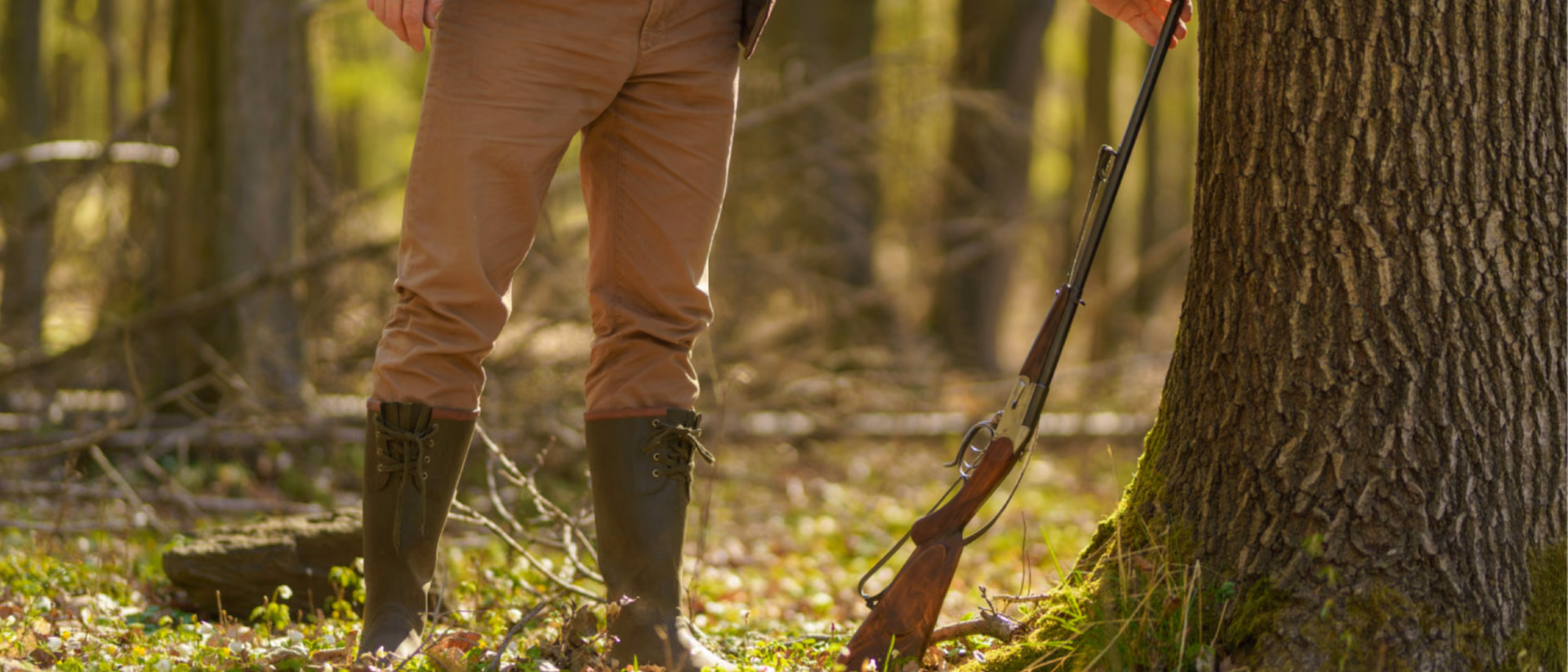 The legs and torso of a hunter standing in a sunlit forest, a broken-open shotgun propped against the tree beside him.