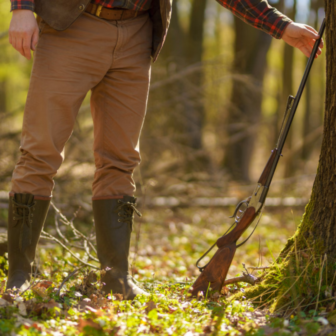 The legs and torso of a hunter standing in a sunlit forest, a broken-open shotgun propped against the tree beside him.