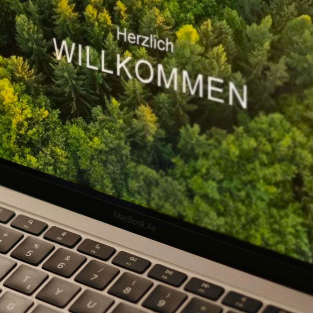 A close-up of a laptop screen greeting the reader with Herzlich Willkommen over an aerial photograph of a forest.