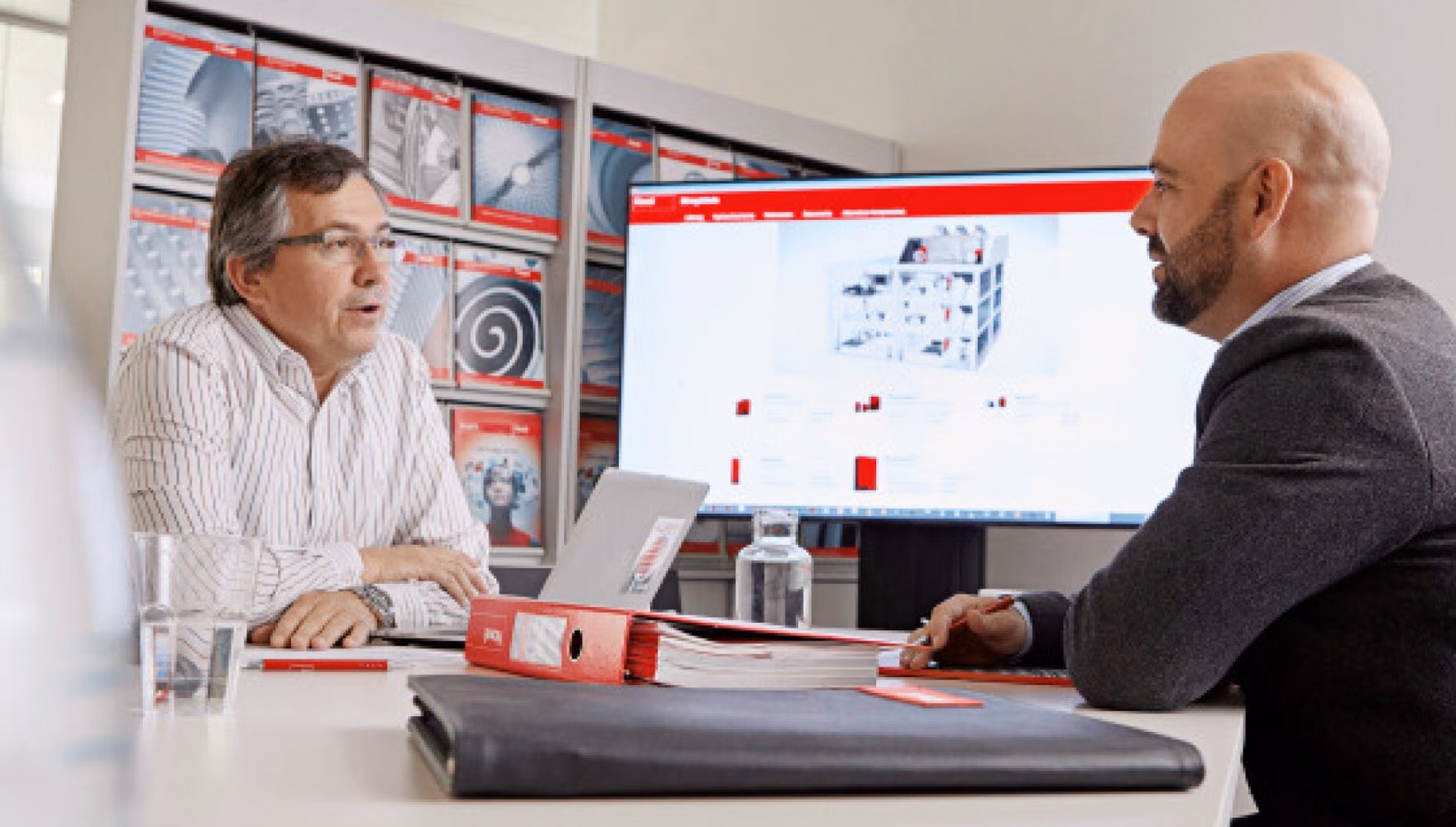Two men sit across a meeting table talking, with a laptop between them and a large screen behind showing a red-branded product page.