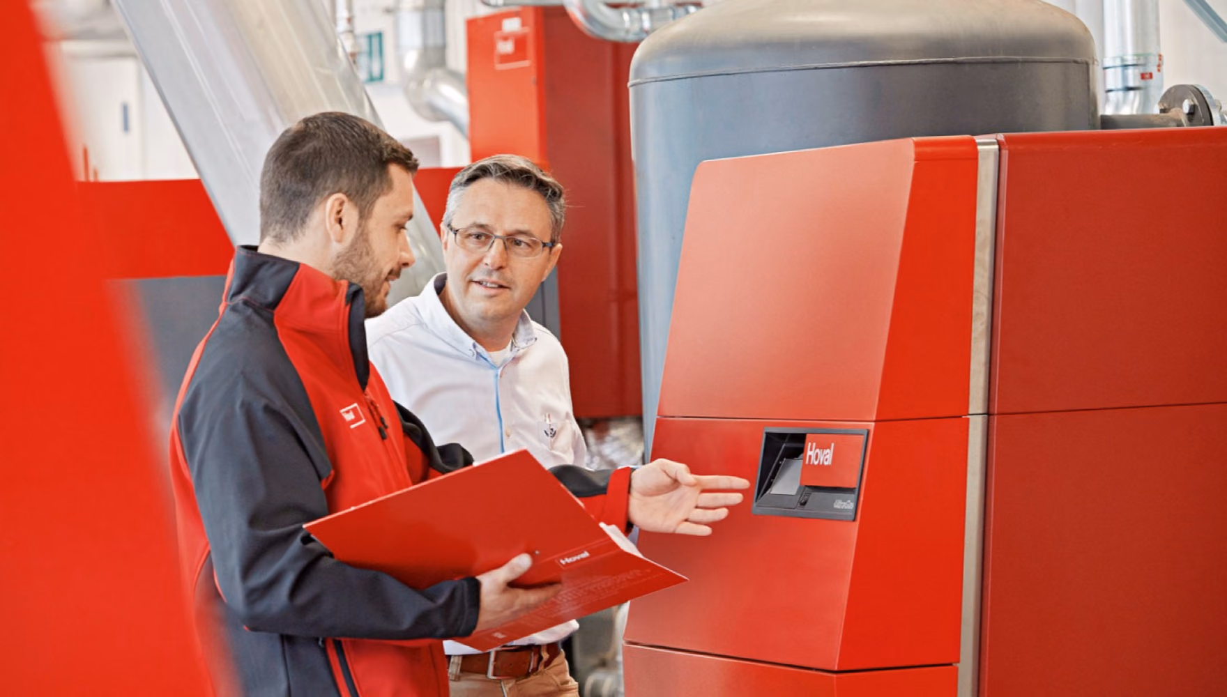 A man in red workwear with an open red folder explains something to a colleague standing beside a red Hoval boiler.
