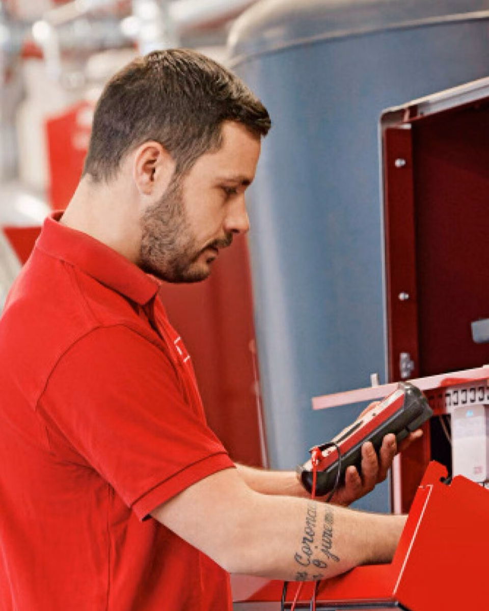 A man in a red polo shirt holds a handheld service device at an opened red heating unit.