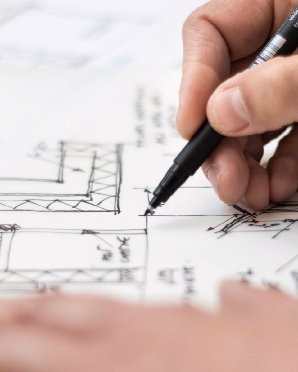A hand draws structural details onto a sheet of hand-drawn plans with a black fineliner.