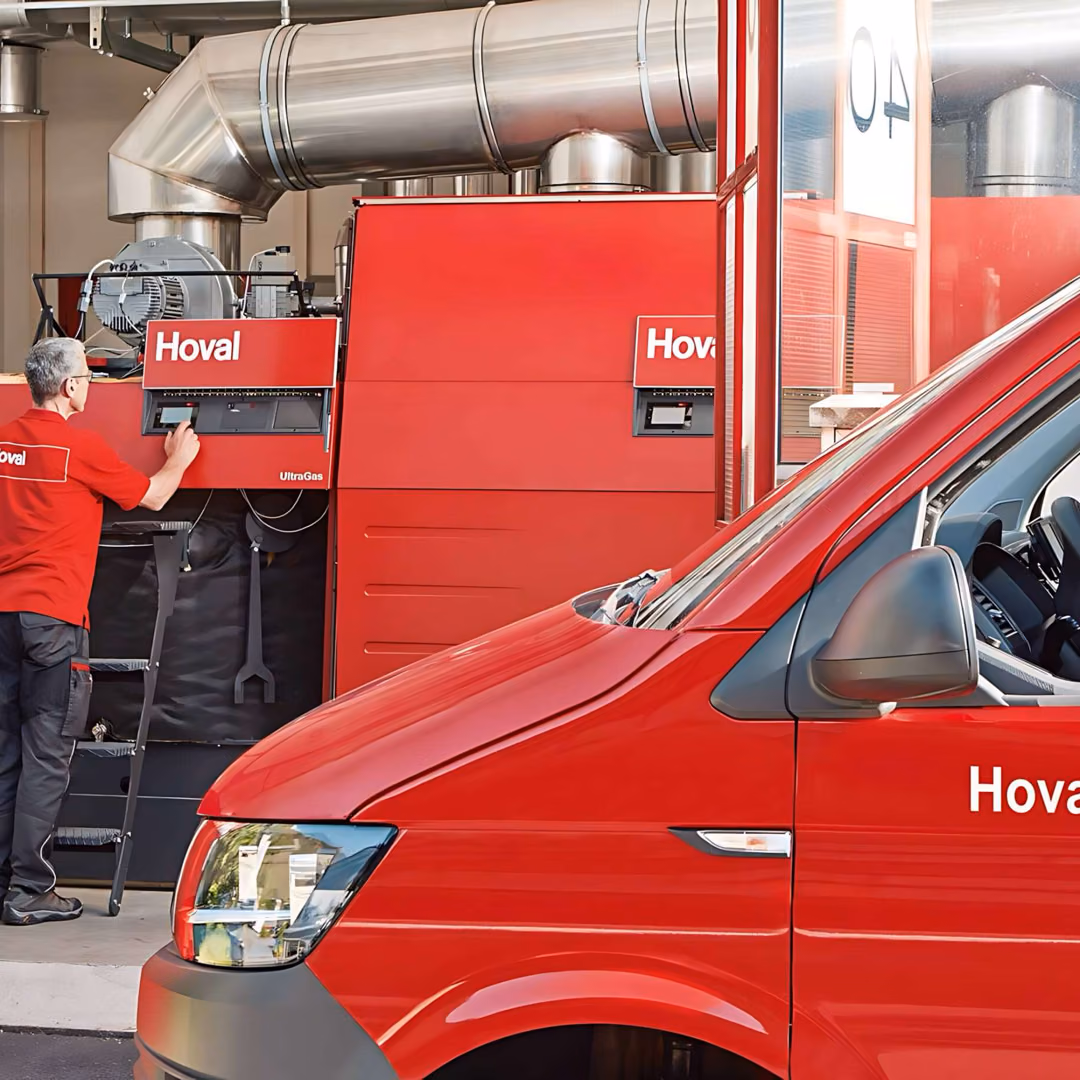 A service technician in a red Hoval polo shirt works at the control flap of a red Hoval UltraGas boiler while a red Hoval.com van stands in the foreground.