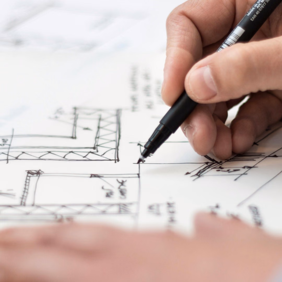 A hand draws structural details onto a sheet of hand-drawn plans with a black fineliner.