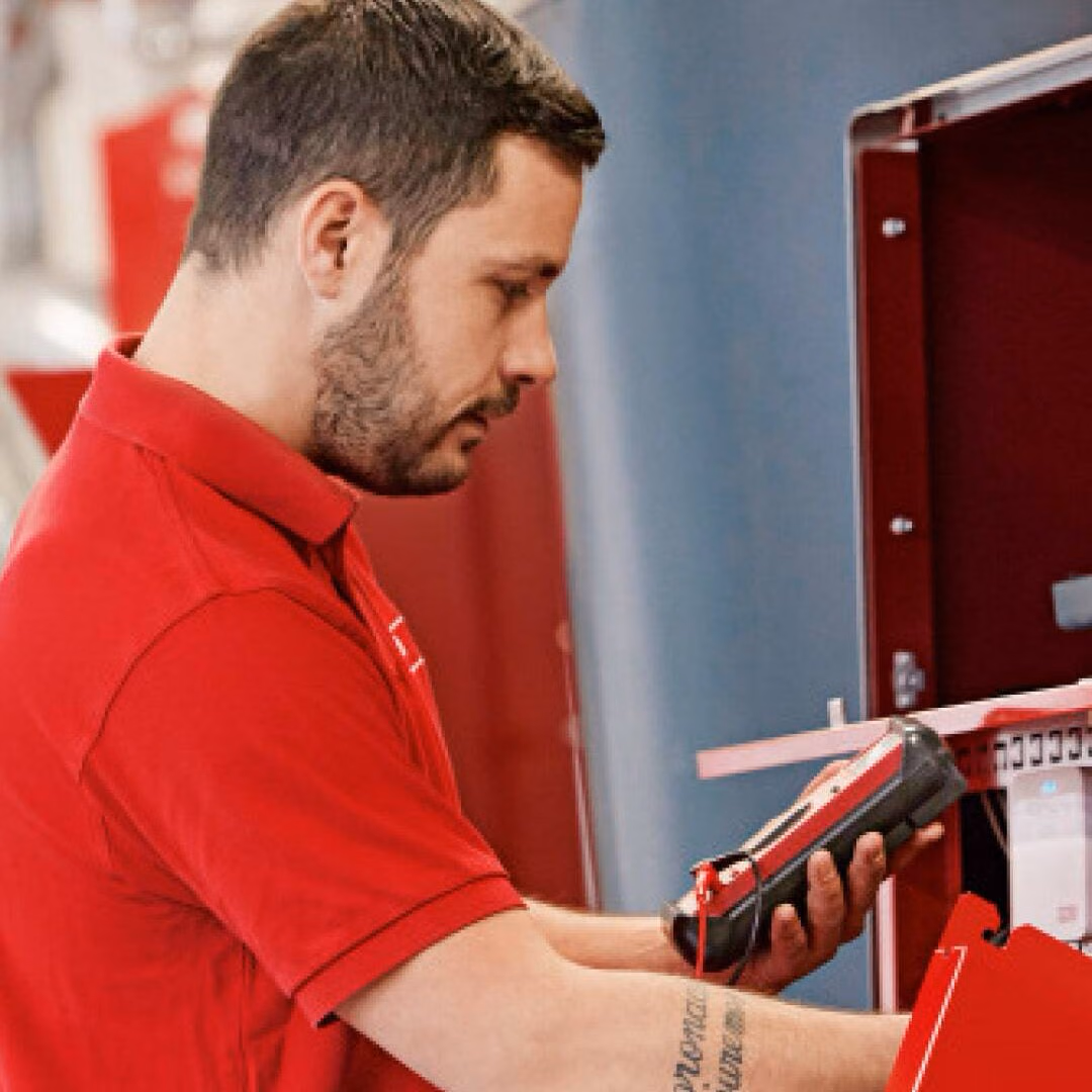 A man in a red polo shirt holds a handheld service device at an opened red heating unit.