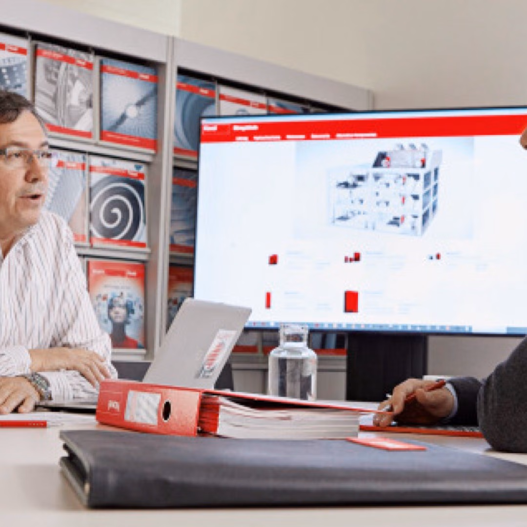 Two men sit across a meeting table in conversation, a laptop between them and a large screen showing a red-branded product page behind.