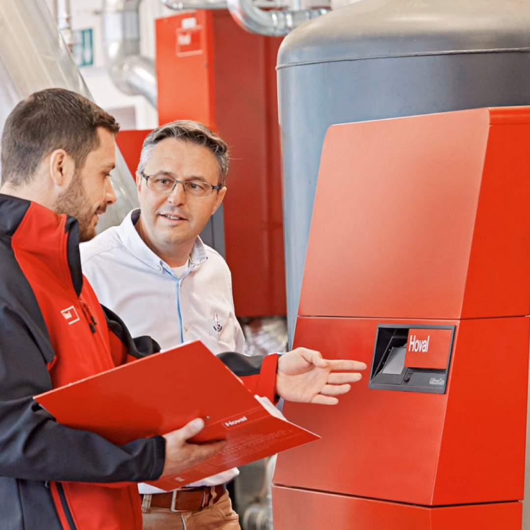 A man in red workwear holding a red folder talks with a colleague in front of a red Hoval boiler in a plant room.