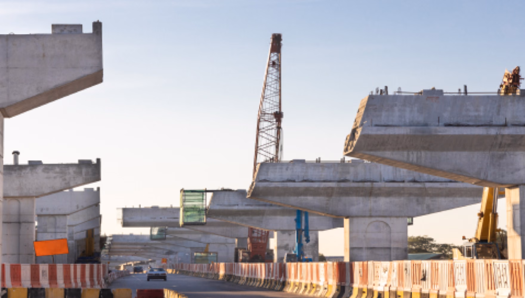 Precast concrete bridge segments and pier caps standing in a line along a road behind orange safety barriers, a lattice crane between them.