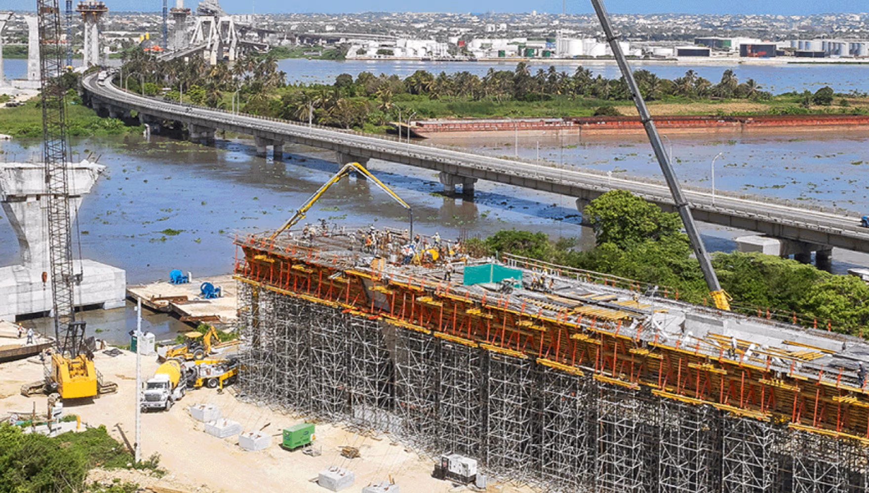 An aerial view of a bridge site beside water, a long wall of red and yellow shoring props under the new deck, a concrete pump boom and a mobile crane at work, the existing bridge running behind it.