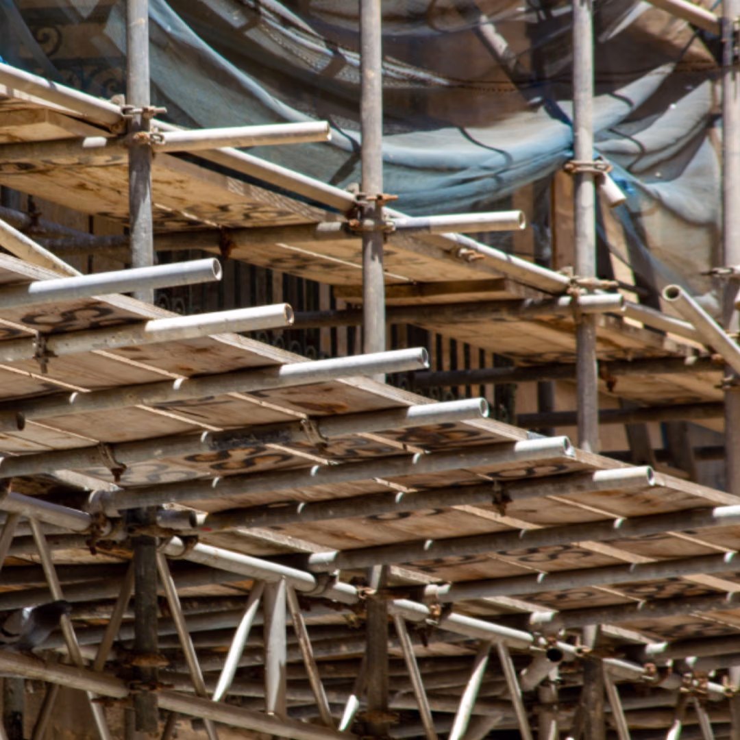 Timber boards and steel tubes of a scaffolding platform stacked in layers, a torn blue safety net behind them.