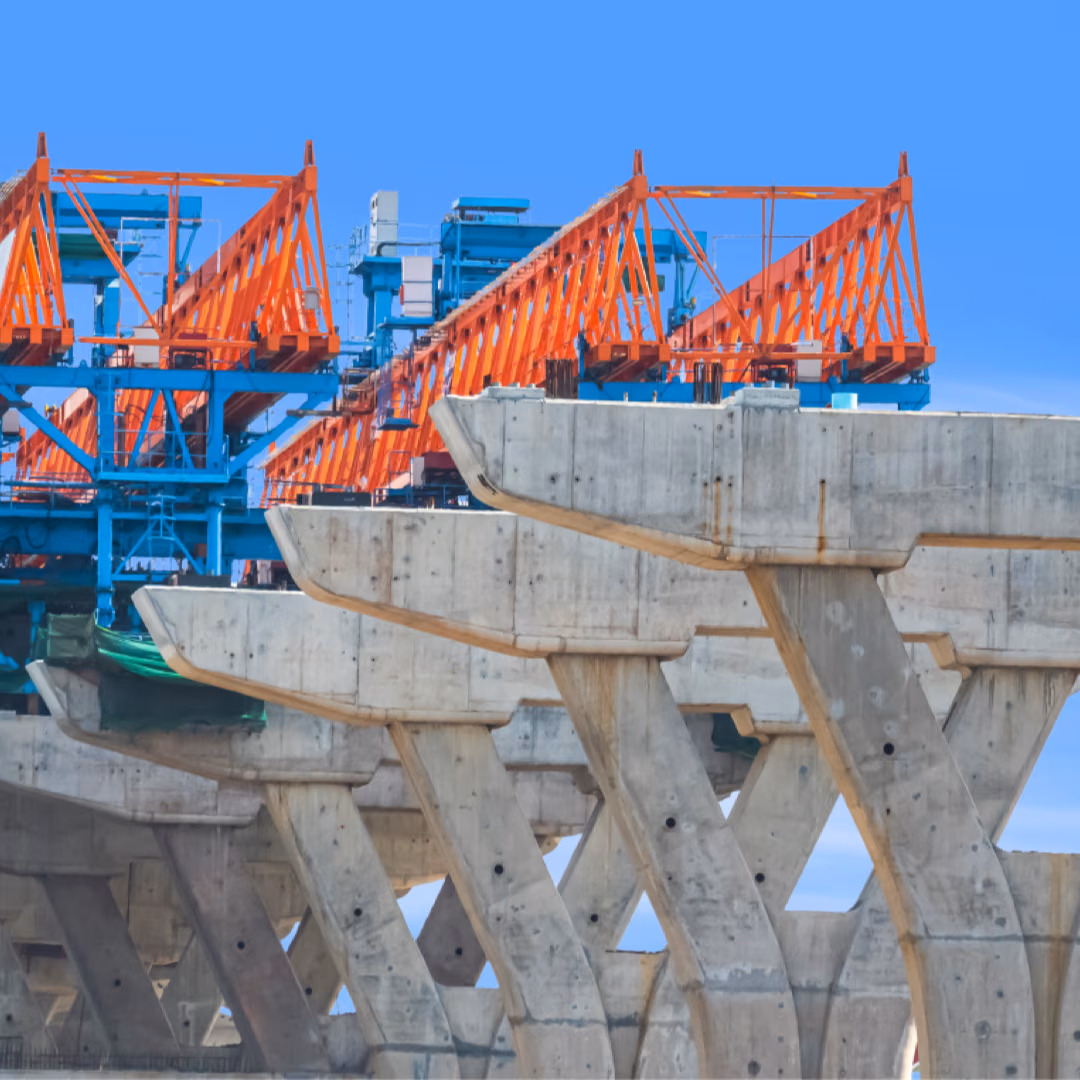 A bridge deck under construction, an orange launching gantry standing on a row of splayed concrete pier caps against a clear blue sky.