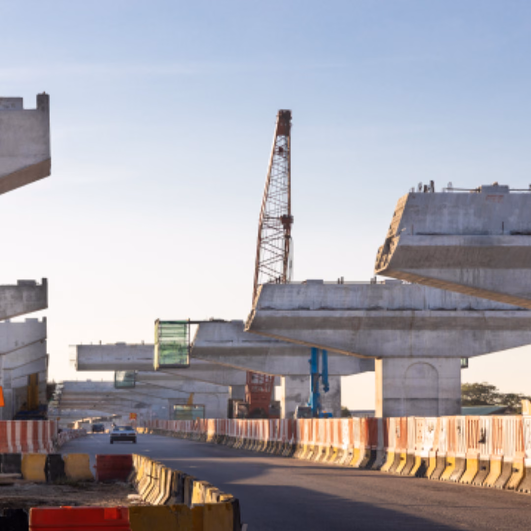 Precast concrete bridge segments and pier caps standing in a line along a road behind orange safety barriers, a lattice crane between them.