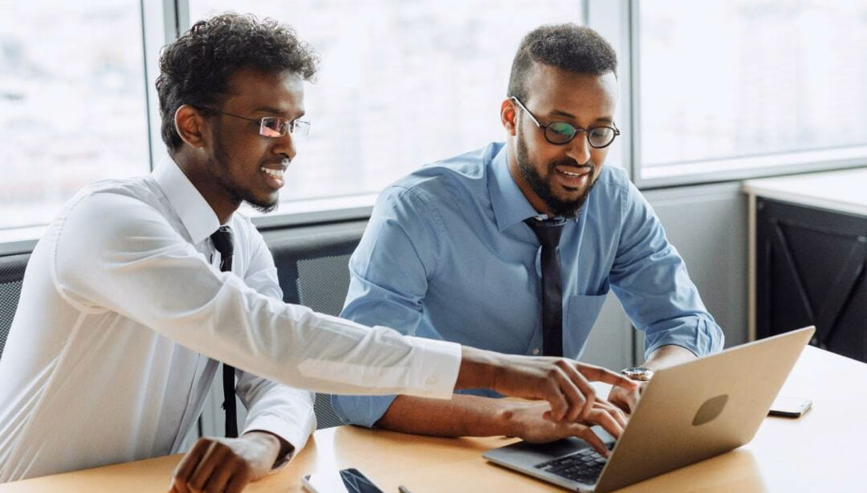Two men in shirts and ties sit at a laptop in a bright meeting room, one of them pointing at the screen.