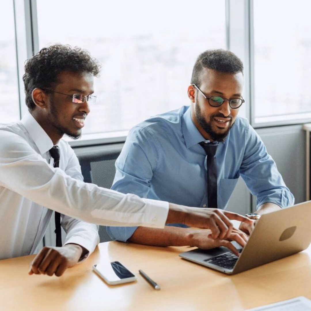 Two men in shirts and ties sit at a laptop in a bright meeting room, one of them pointing at the screen.