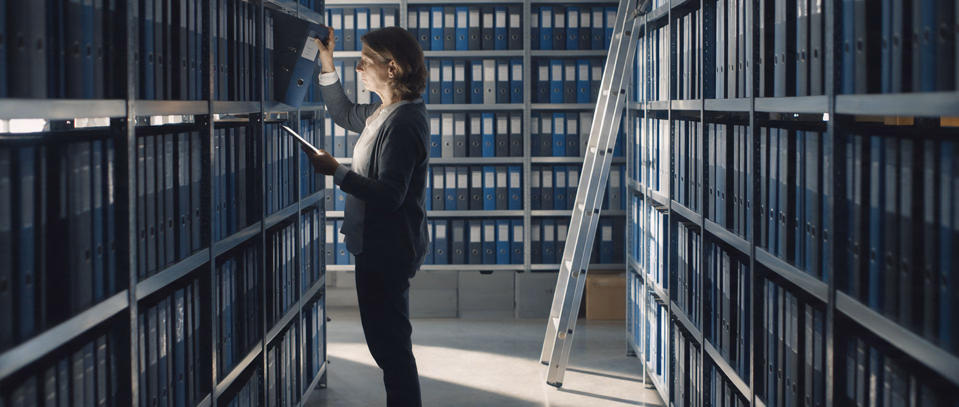A woman in a grey cardigan tilts a binder out of a shelf in a long archive aisle of dark blue binders while holding a tablet, a rolling ladder leaning against the shelving behind her.