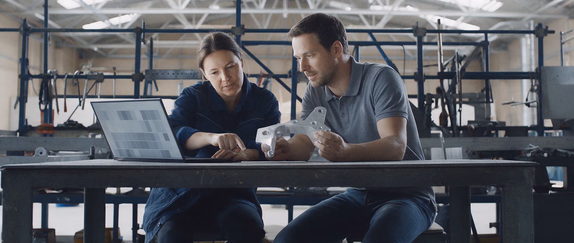 Two colleagues sit at a workbench in a bright assembly hall, one pointing at a laptop screen while the other holds a small machined aluminium bracket.