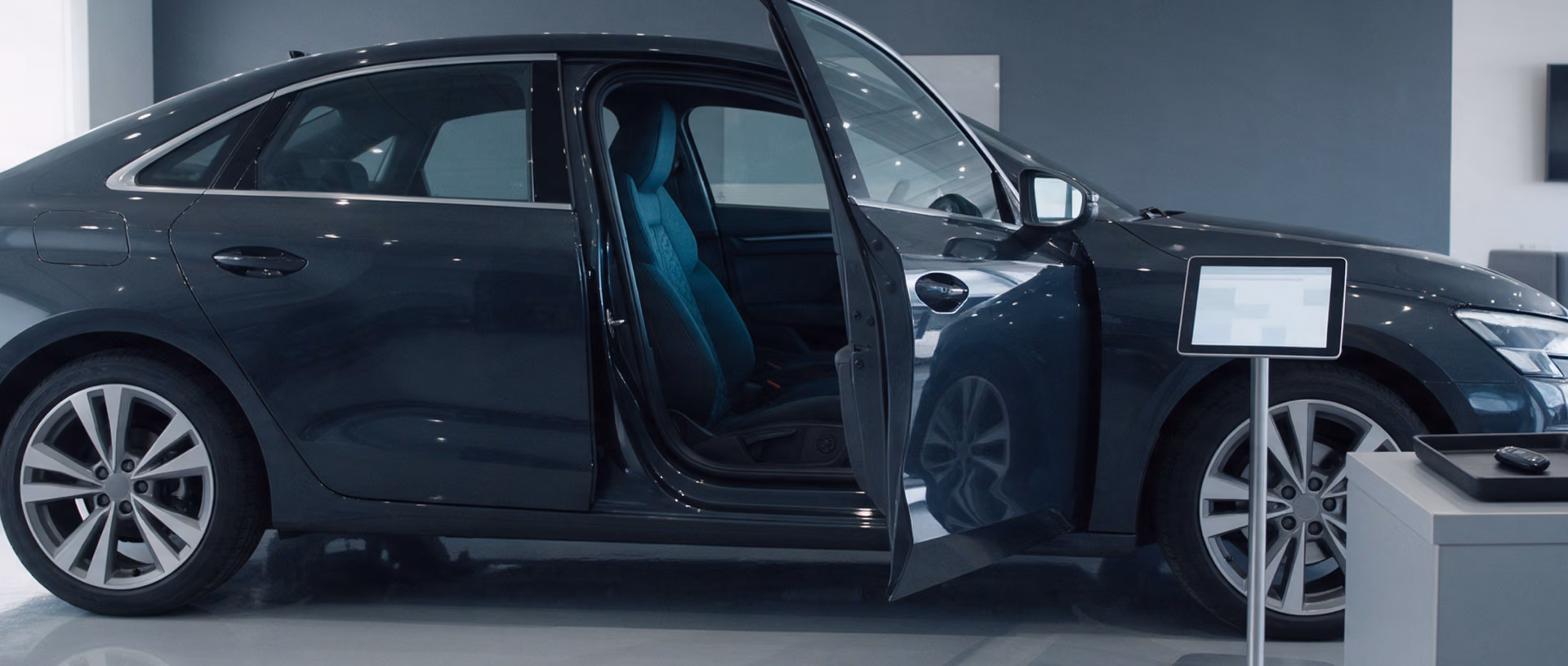 A dark grey car with its driver door open stands in an indoor handover bay beside a tablet on a floor stand and a key tray, a teal seat visible through the door.