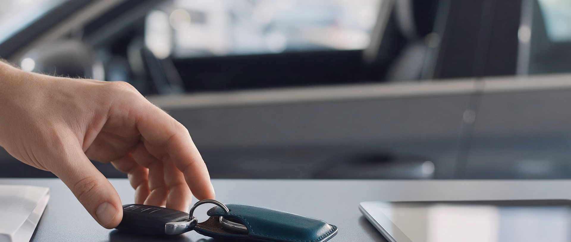 A hand places a car key beside a teal leather key wallet on a dark handover desk, with a closed folder to the left, a tablet to the right and a car door behind.