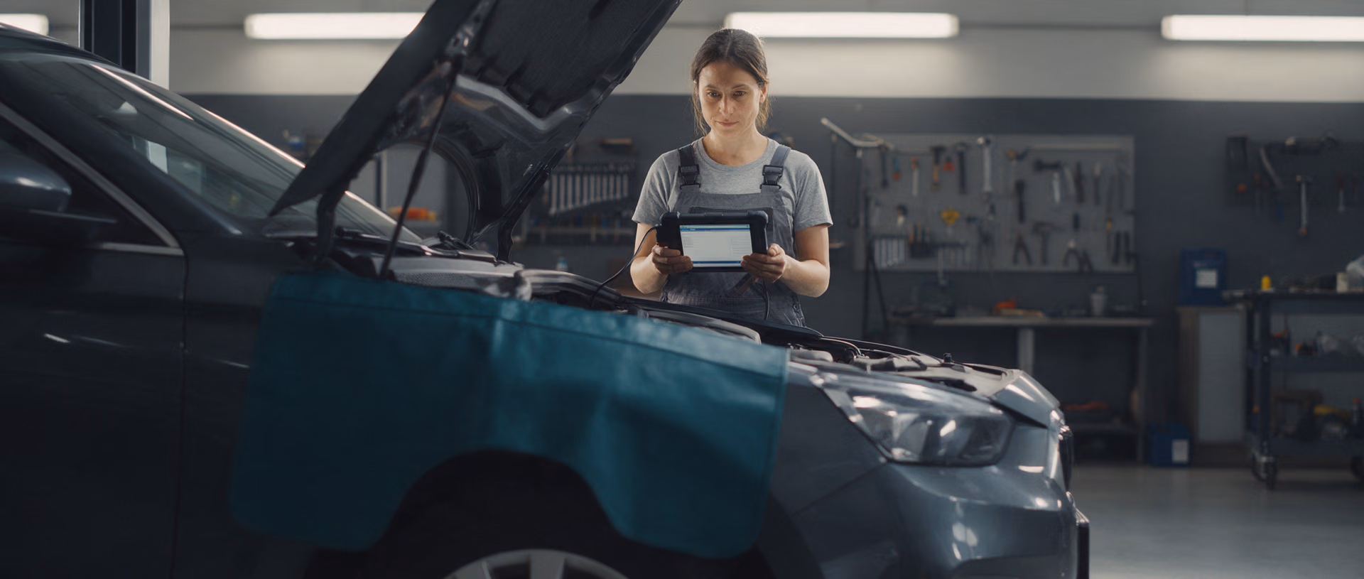 A mechanic stands at the open bonnet of a dark grey car in a workshop at night, reading a diagnostic tablet connected to the engine bay, a teal fender cover over the wing.