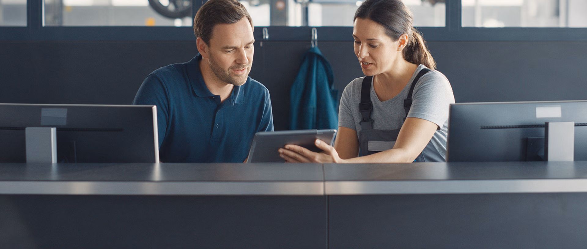 A service advisor and a mechanic sit at a dark reception desk with two monitors, looking at a tablet, a teal workshop jacket hanging on the wall behind them.