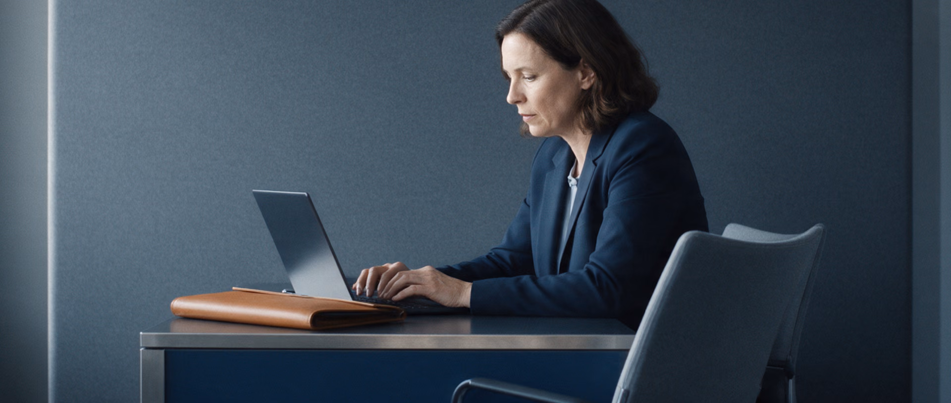 A woman in a dark blazer types notes on a laptop at a bank advisory desk, a closed tan leather folder beside her and the chair opposite empty.