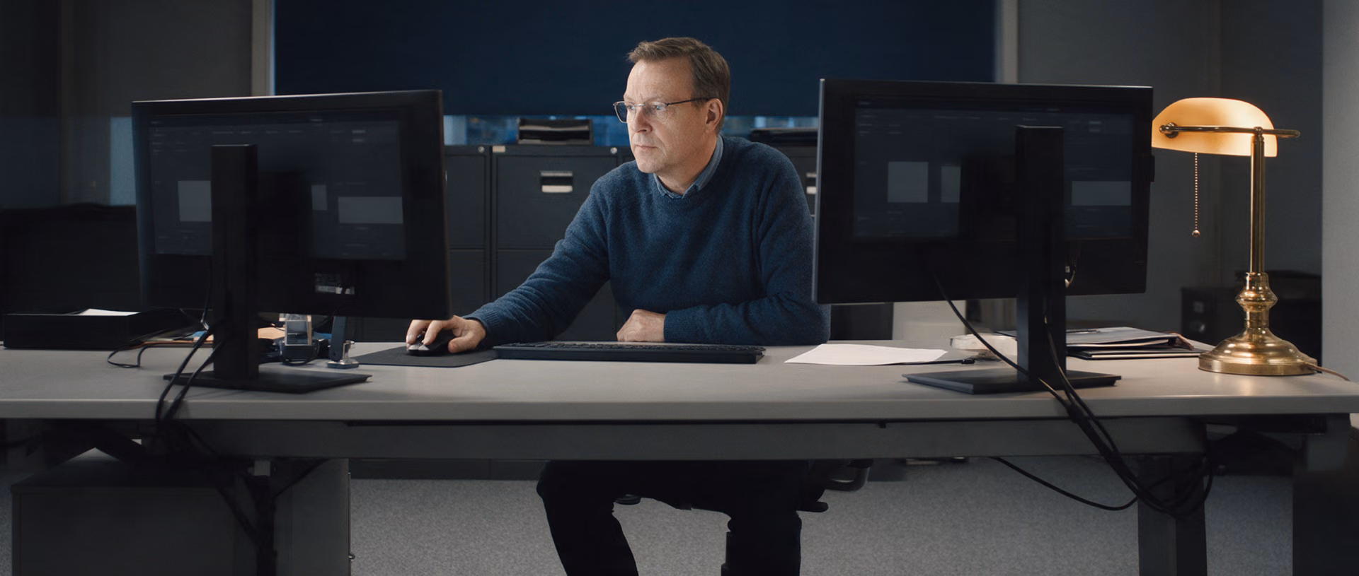 A man in a dark blue jumper and glasses compares two dark monitors at an office desk in the evening, a brass lamp lit at the right end of the desk.