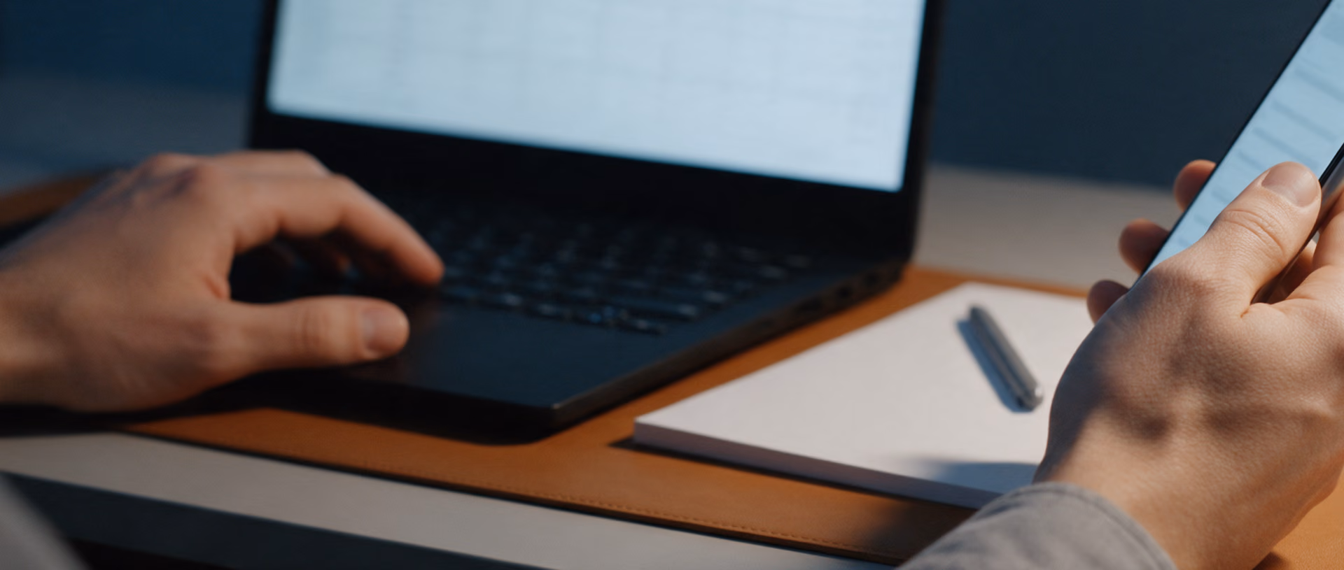 Close-up of a man's hands at a desk at dusk, one on a laptop showing a spreadsheet grid, the other holding a smartphone, a blank notepad and a pen on a tan leather desk mat.