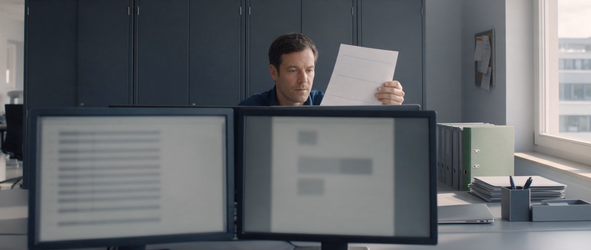 A man in a dark blue shirt holds a printed sheet up beside one of two monitors in an office with dark cabinets, a green binder standing in a stack on the desk.