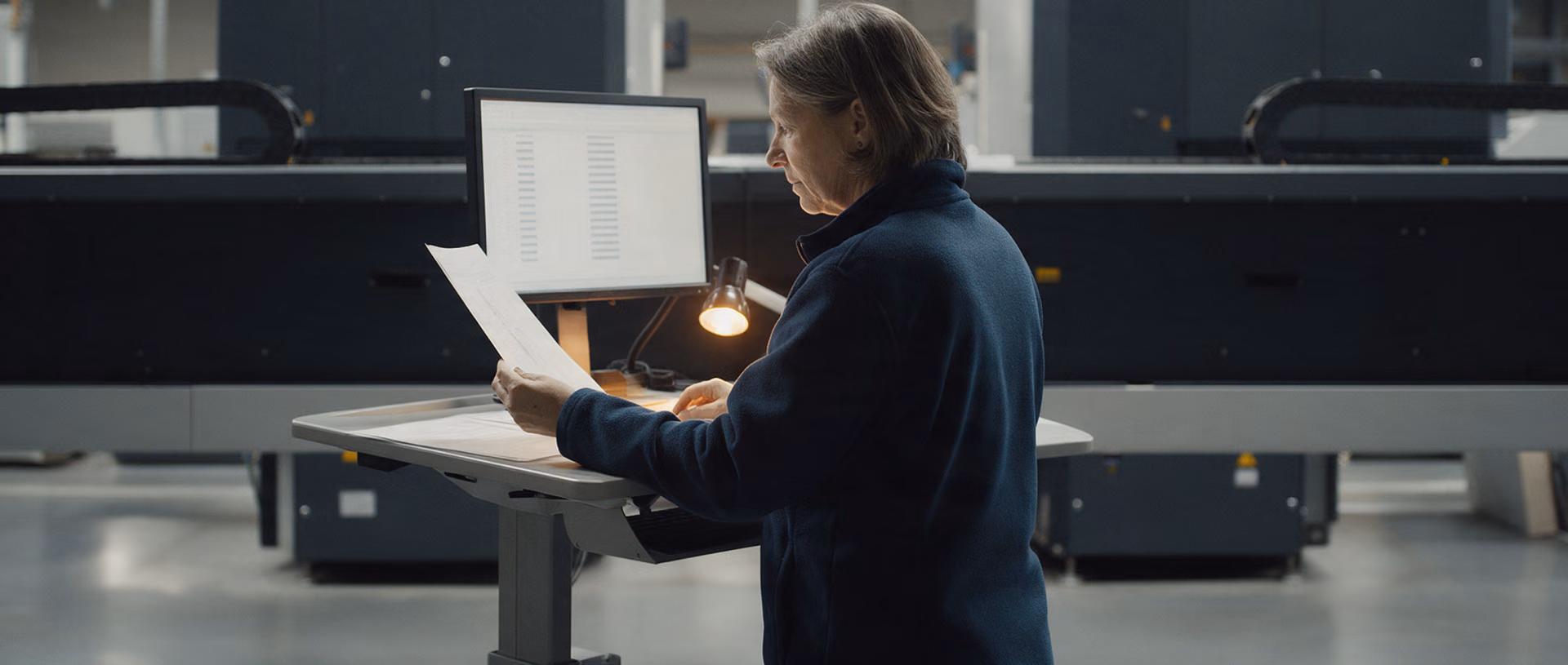 A production planner stands at a standing desk at the edge of a production hall at night, comparing a sheet of paper with a monitor under a small work lamp.