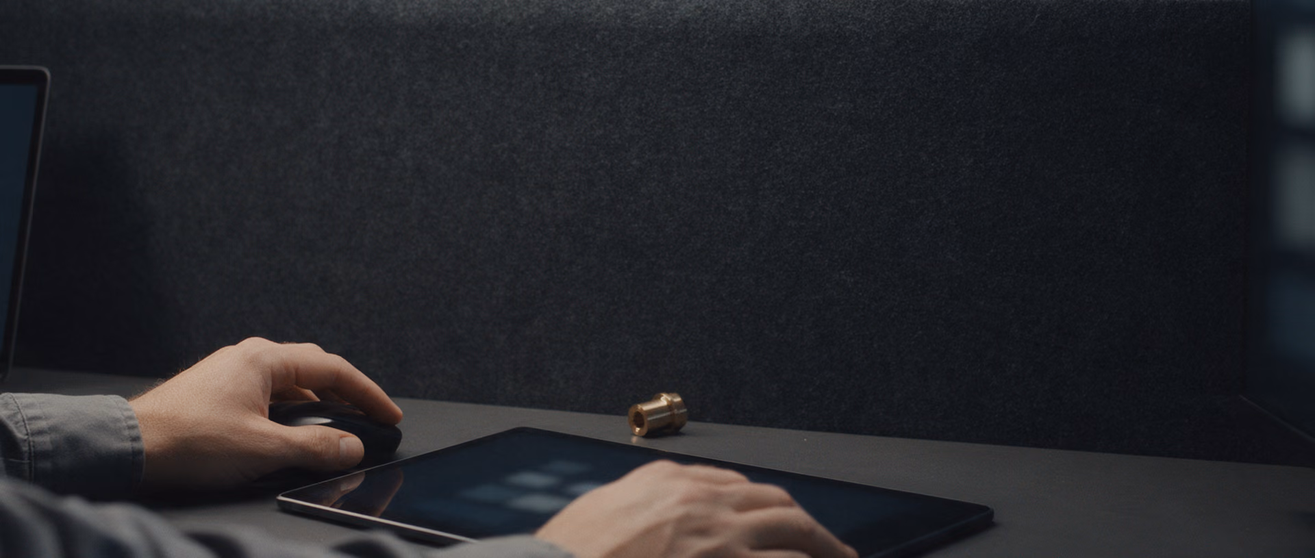 Hands rest on a mouse and a tablet on a dark desk in front of a grey felt partition, with a laptop and a monitor at either edge and a small brass part between them.