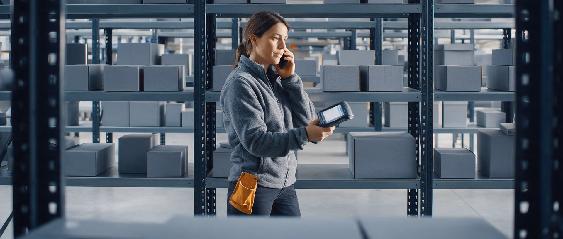A warehouse worker in a grey fleece stands between tall shelves of grey boxes, phone at her ear and a handheld scanner in her hand, a leather tool pouch on her belt.