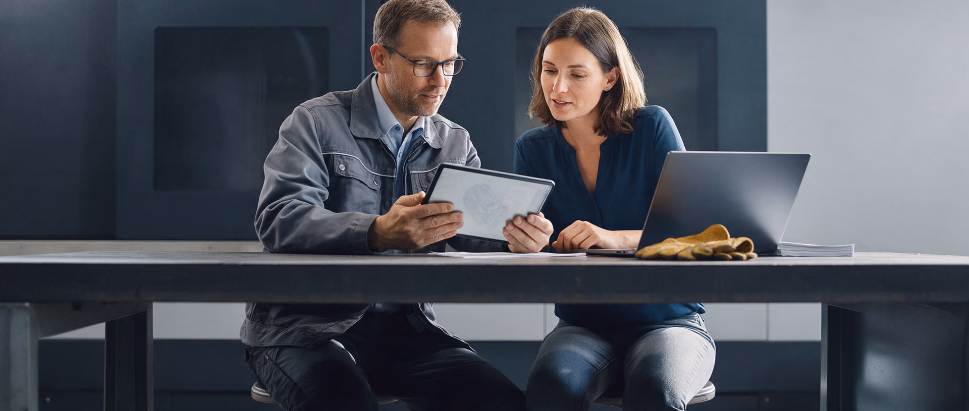 An engineer and a sales colleague sit at a steel workbench in a machine hall, looking at a tablet beside an open laptop and a worn leather work glove.