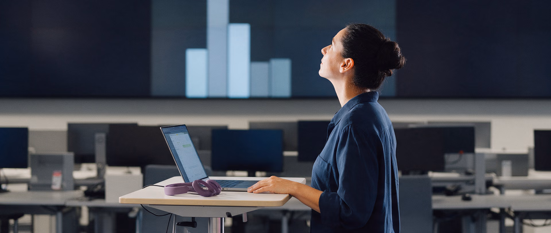 A woman at a standing desk in a service centre looks up at a large wall screen showing blurred bars, a violet headset lying beside her laptop.