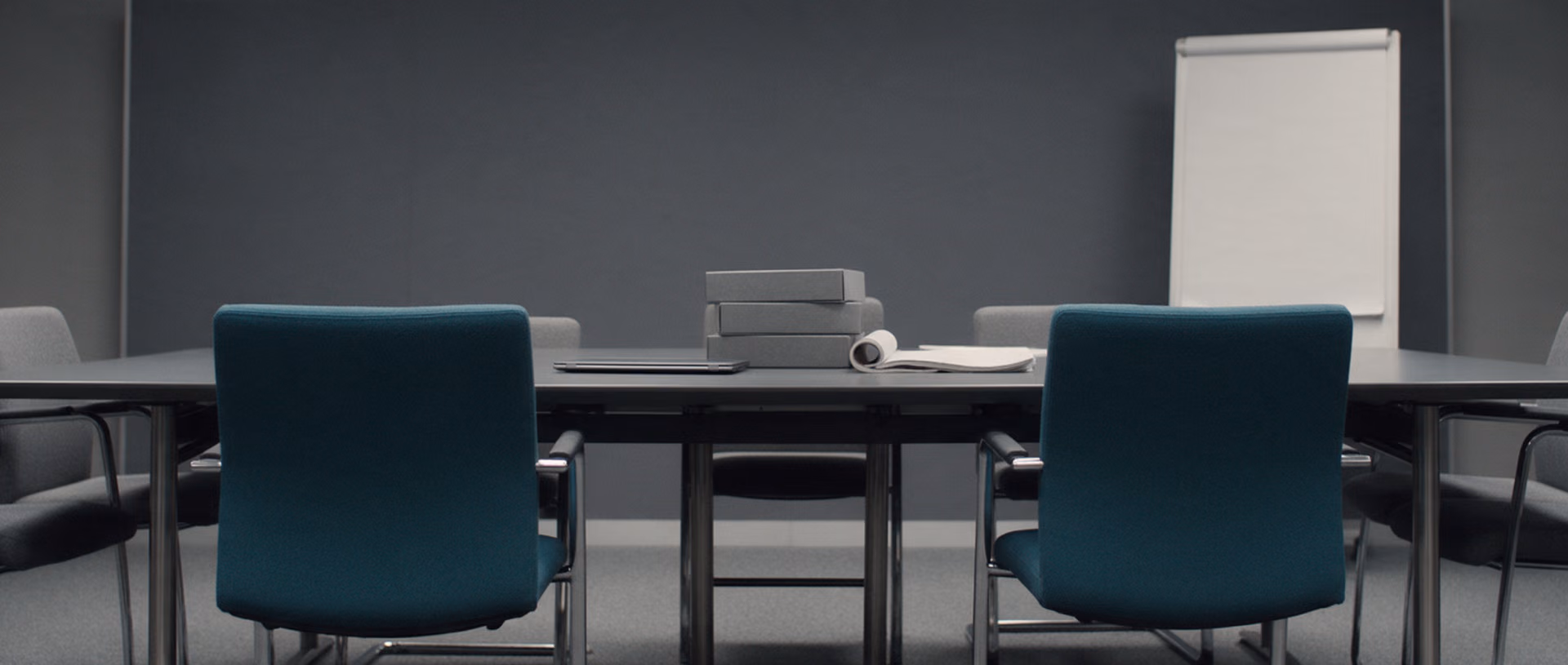 An empty meeting room in the evening with a closed laptop, grey archive boxes and a rolled flipchart pad on a long table, two teal chairs pushed in.