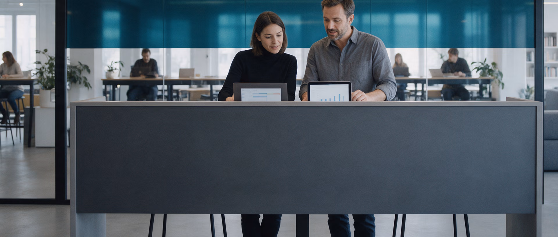 Two consultants stand at a high table in a coworking space, one with a laptop and one with a tablet, a band of teal frosted film on the glass wall behind them.